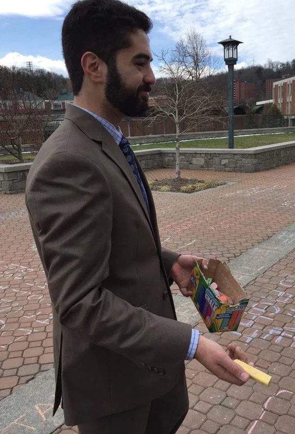 Grant in a suit holding a box of sidewalk chalk on the App State campus